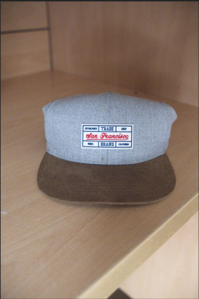 Original Fujifilm RAW photo of a gray and brown baseball cap with a San Francisco Trade patch, resting on a wooden shelf, before noise reduction