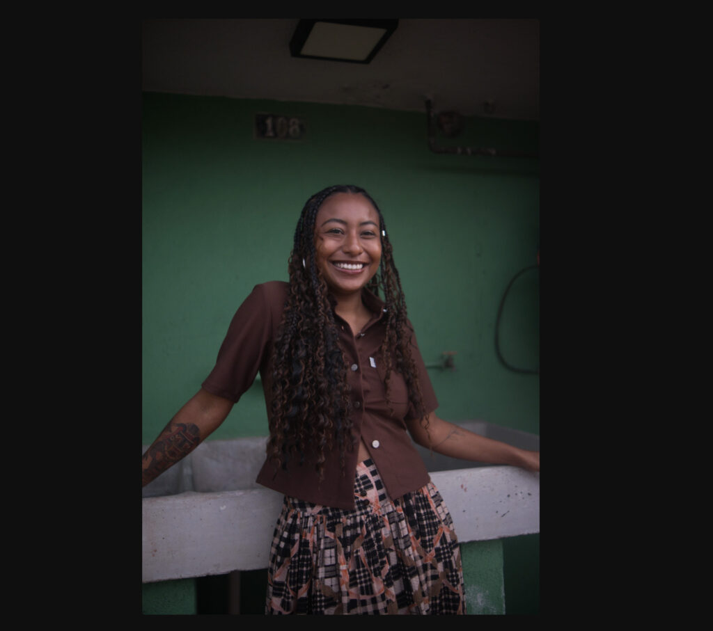 Original RAW portrait of a smiling woman with long braided hair against a green wall, with distracting objects in the background before object removal